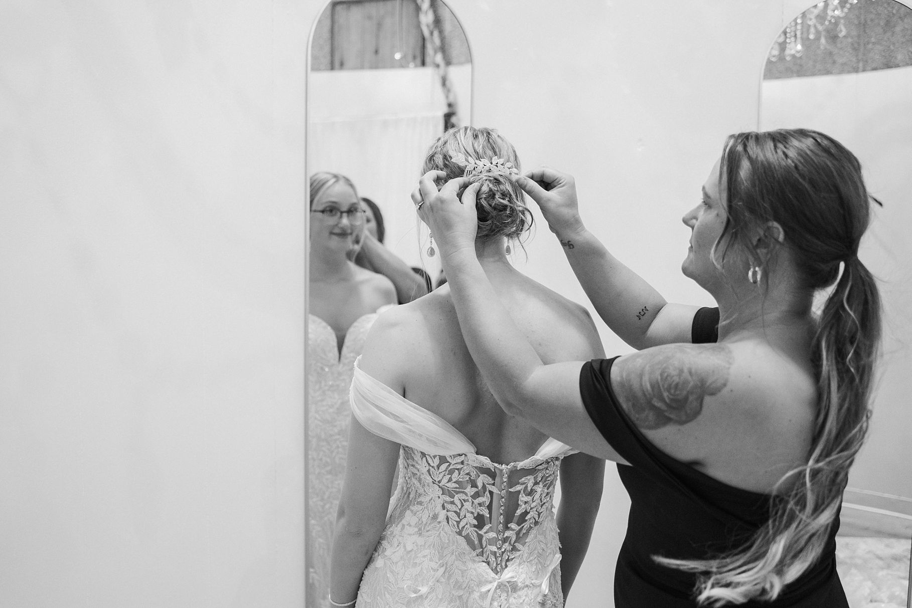 Black and white photo of tattooed bridesmaid adjusting brides updo hairstyle in lace wedding gown by mirror.