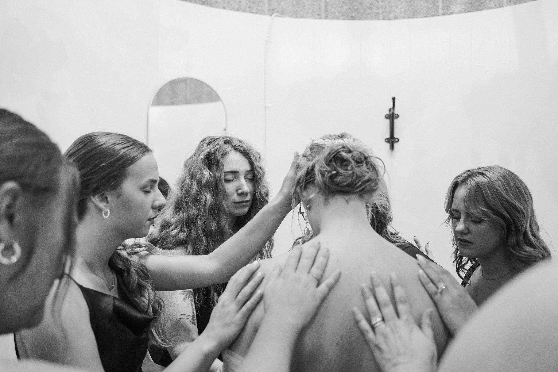 Black and white photo of bridesmaids lovingly placing hands on brides back in a touching pre-wedding moment.