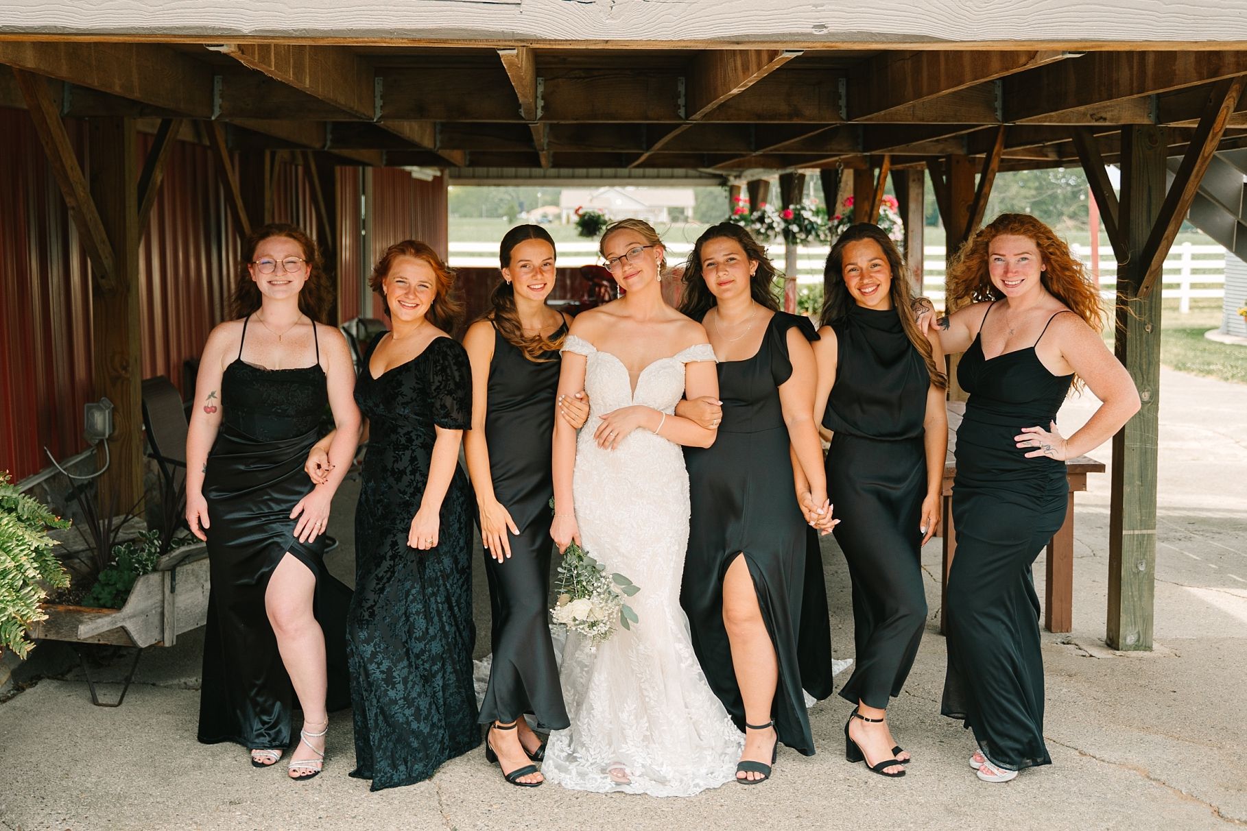 Bride in lace wedding gown poses with six bridesmaids in black dresses under rustic wooden venue structure.