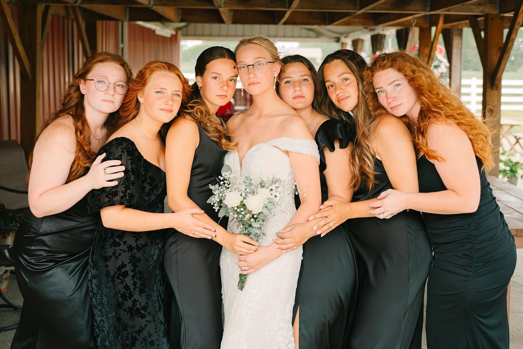 Bride holding white bouquet surrounded by smiling bridesmaids in black gowns in a warm group wedding photo.