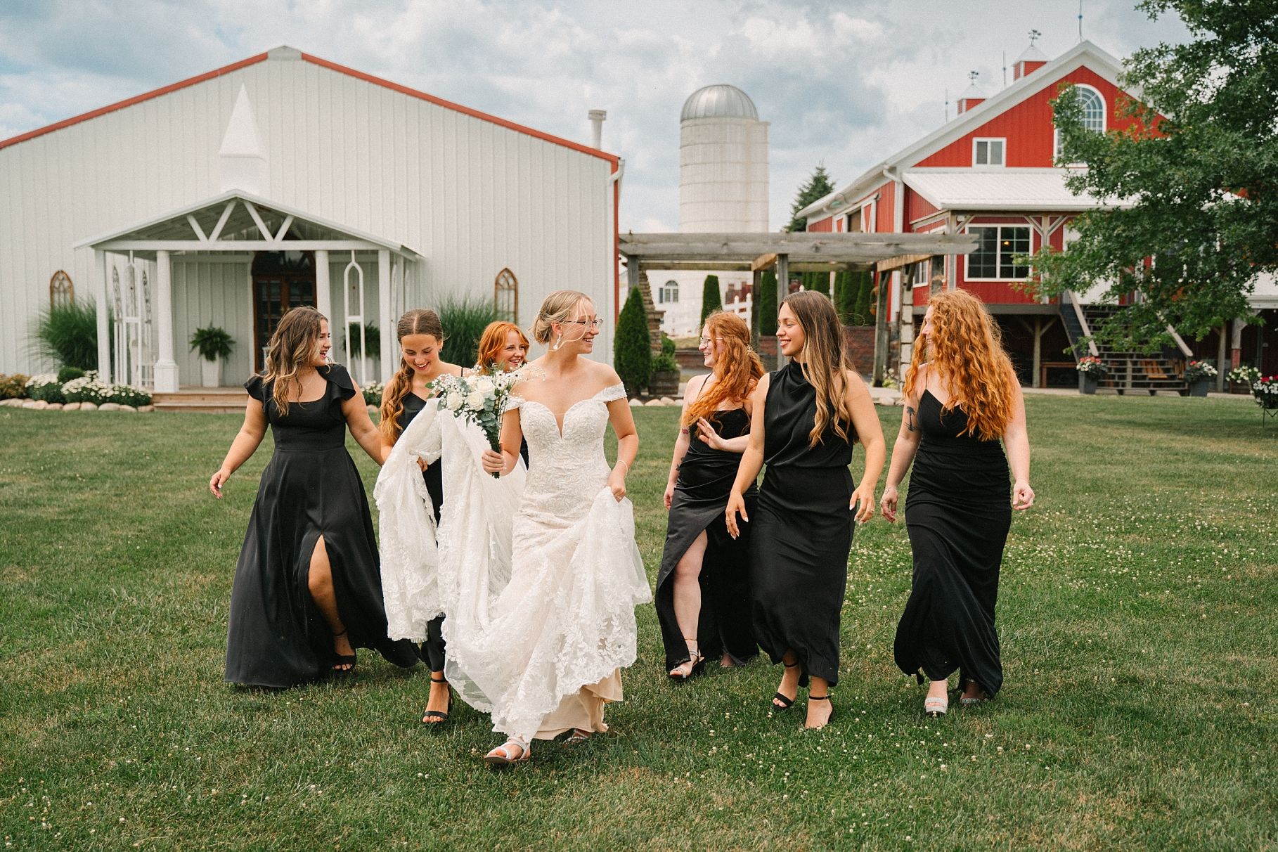 Bride in flowing lace gown walks joyfully with bridesmaids in black dresses across green lawn near white barn.