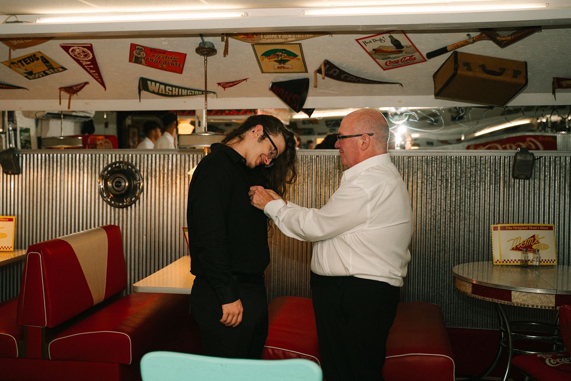 Older gentleman pinning boutonniere on person in black outfit inside retro diner with vintage decor.