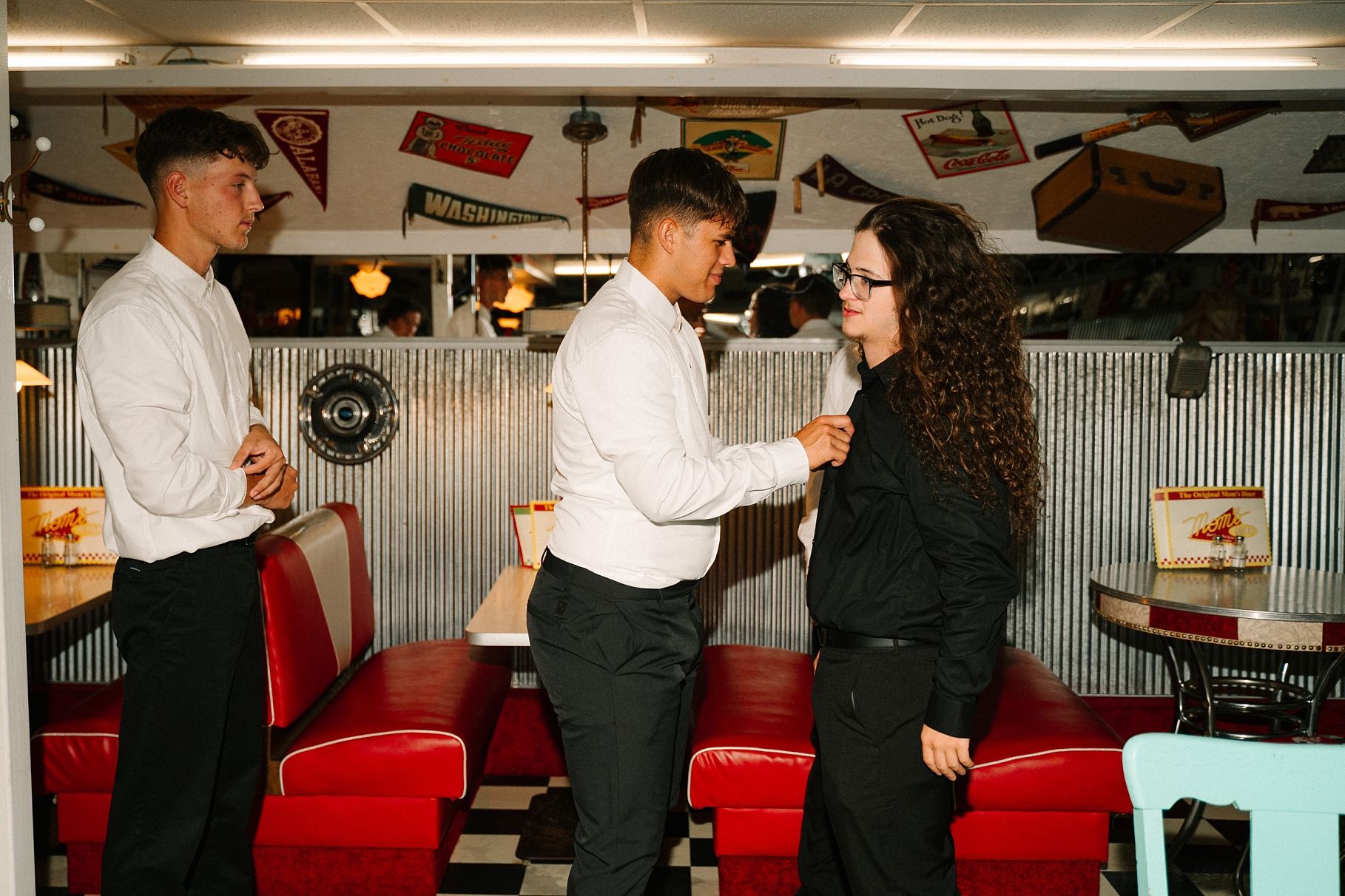 Groomsman in white shirt pinning boutonniere on bridesmaid in black inside a vintage retro-style diner venue.