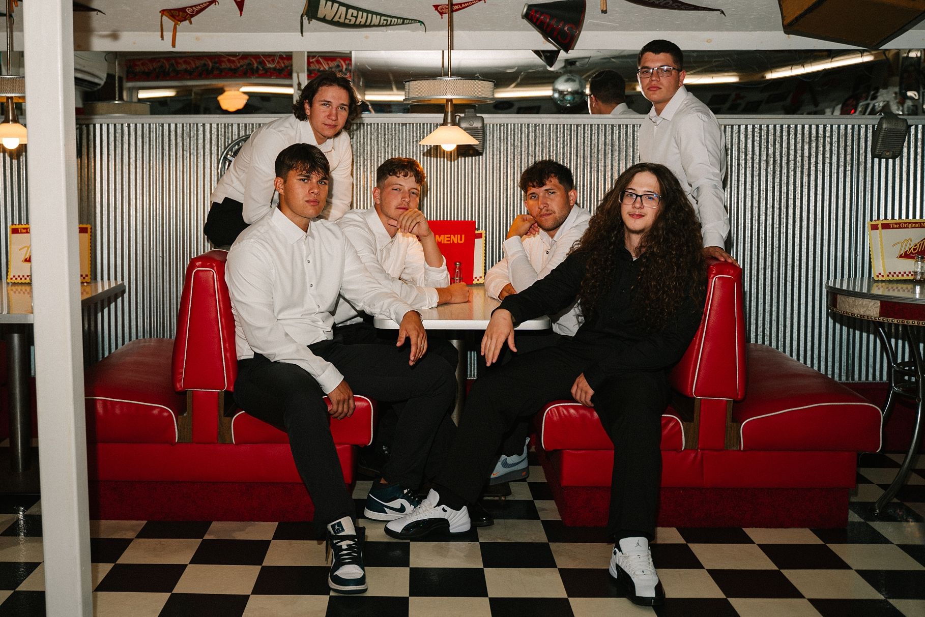 Six young men pose in a retro diner with red booths and checkered floors.