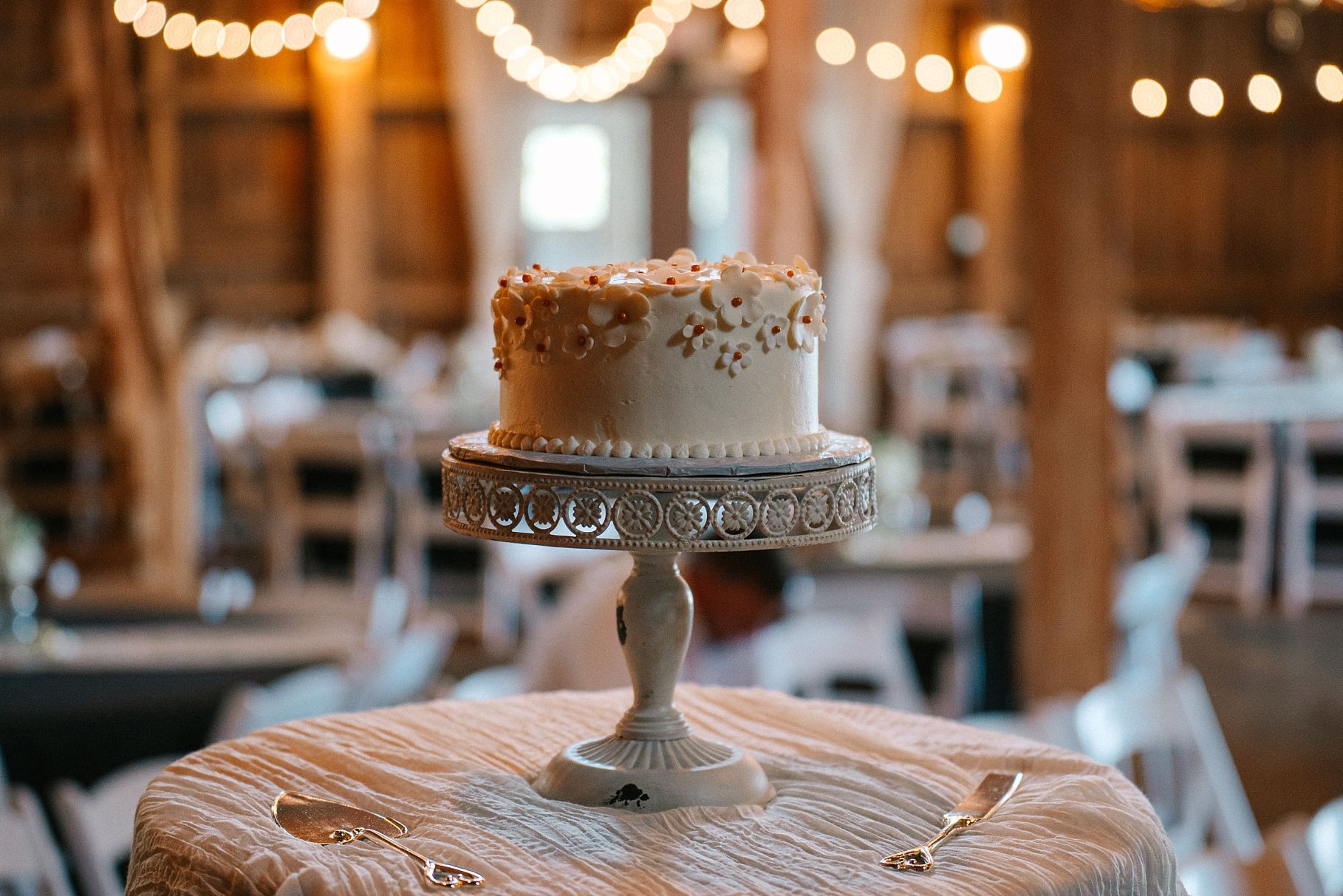 Single-tier white wedding cake with gold details displayed on ornate silver stand in barn venue.