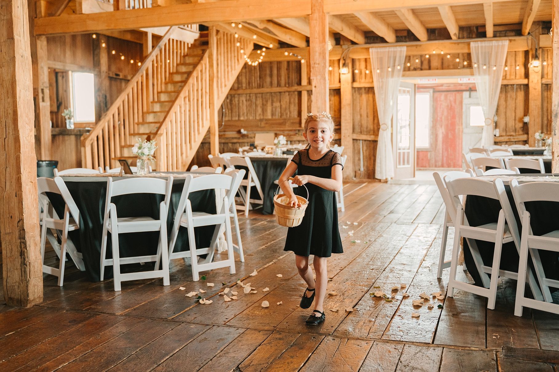 Young flower girl scatters petals across rustic barn wedding venue with wooden floors.