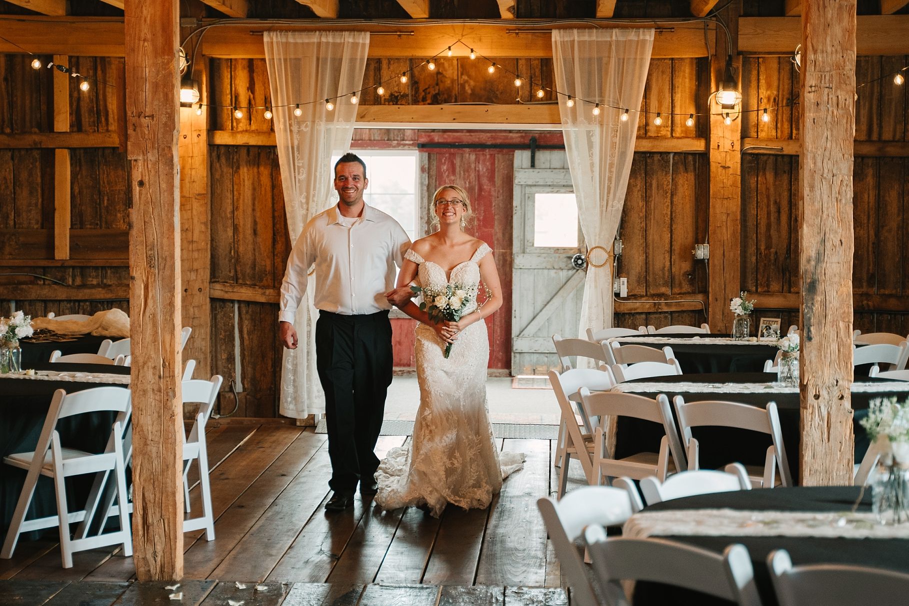 Bride in lace gown walks down aisle with groom in rustic barn wedding venue.