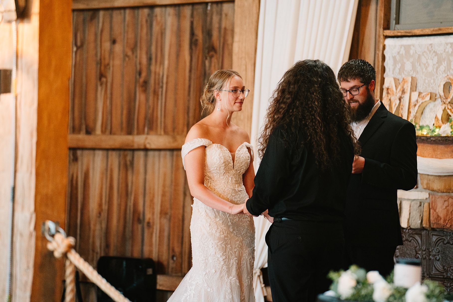 Bride in lace wedding gown exchanging vows with partner during rustic barn wedding ceremony.