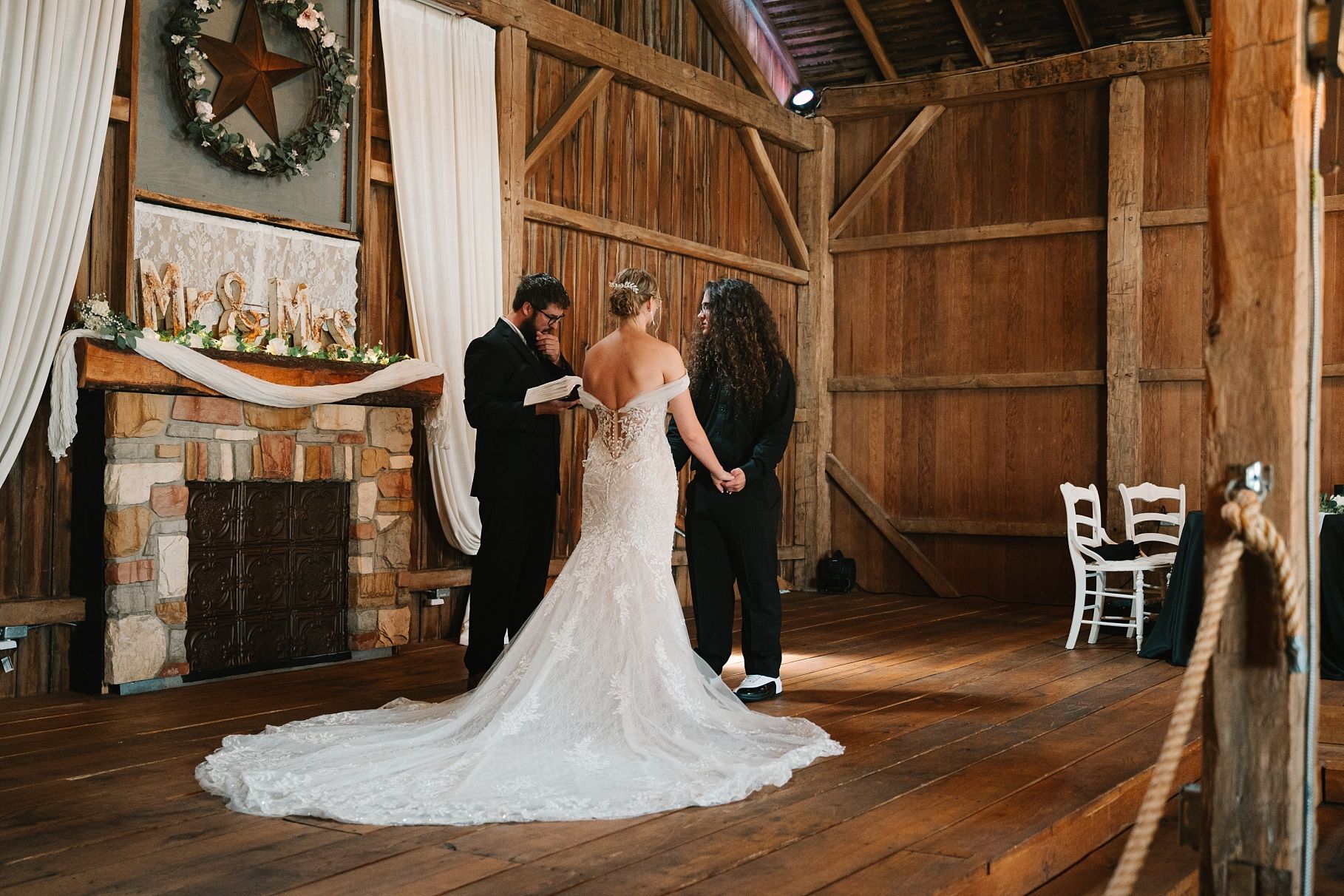 Bride in elegant lace gown stands at rustic barn altar with officiant during ceremony.