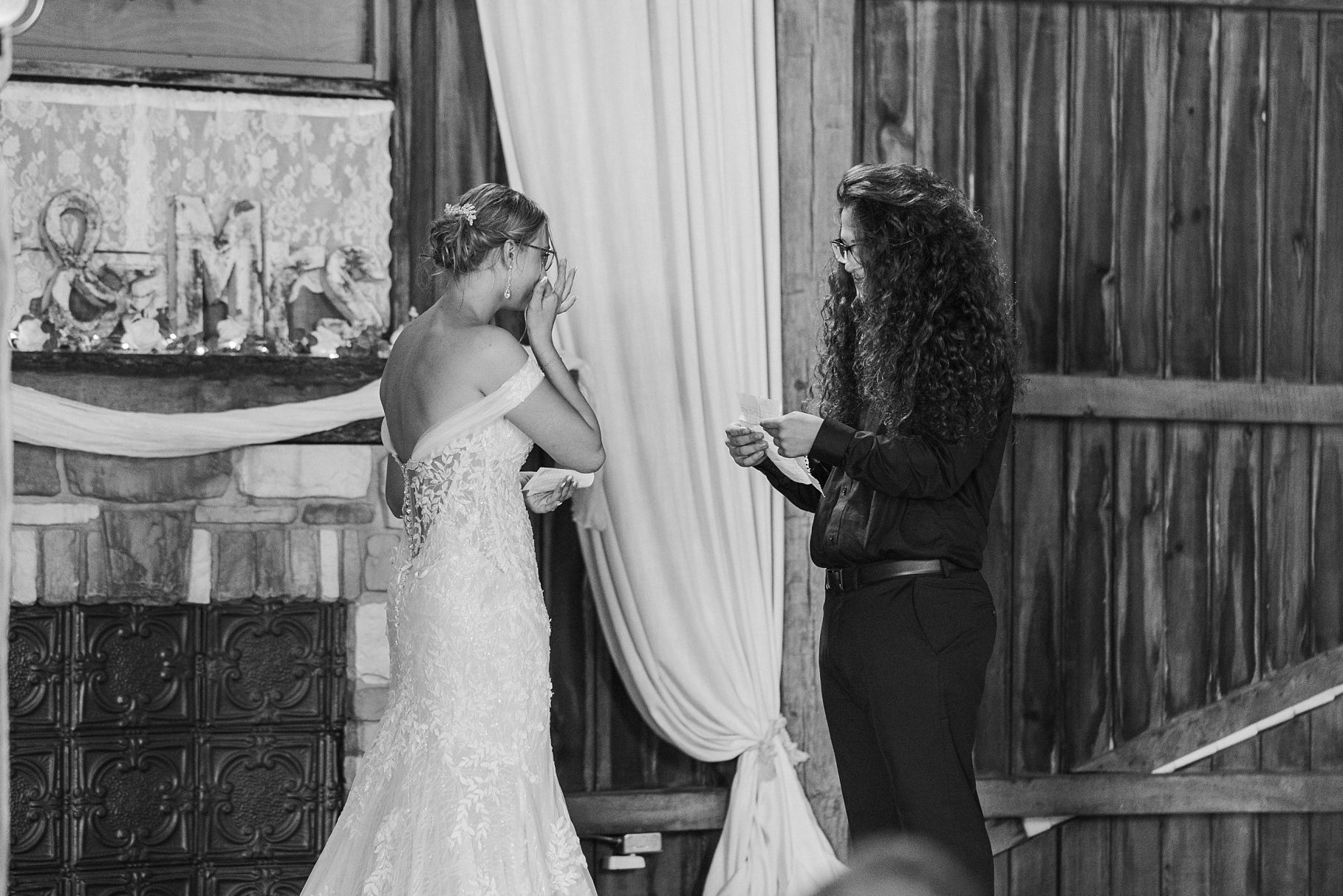 Black and white photo of bride in lace gown sharing emotional moment during barn wedding ceremony.