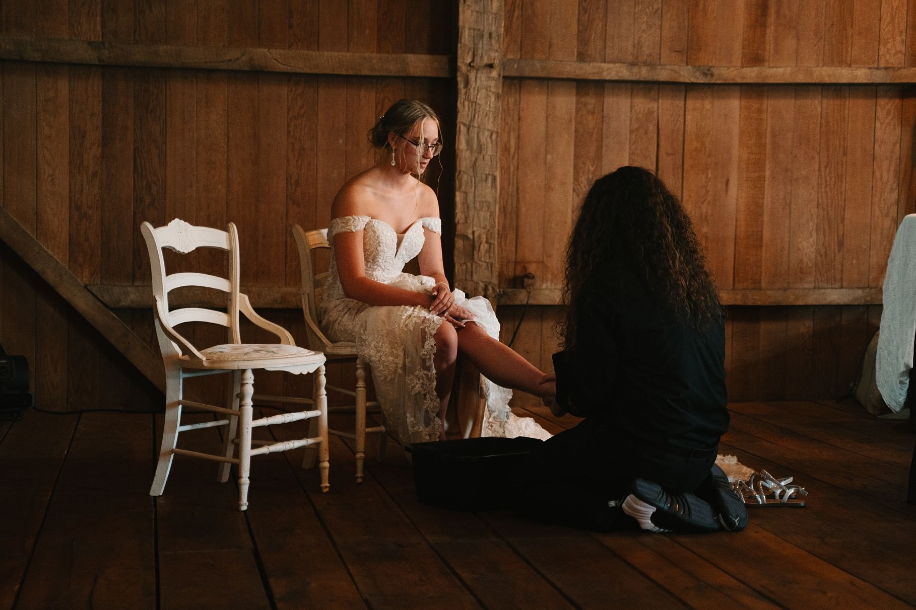 Bride in off-shoulder lace gown seated on chair while partner sits on barn floor during wedding.