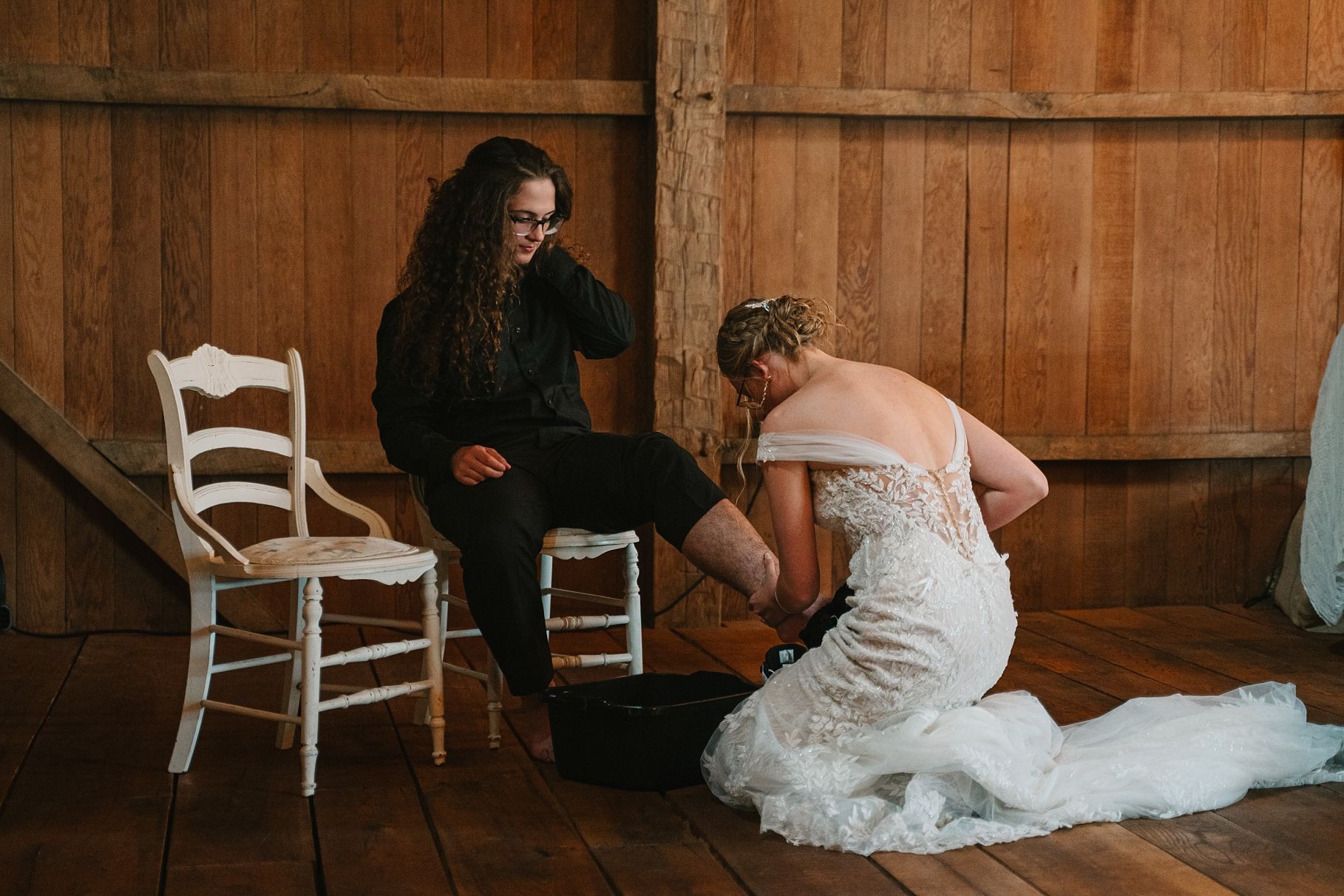 Bride in lace wedding dress performing foot washing ceremony for partner in rustic barn setting.