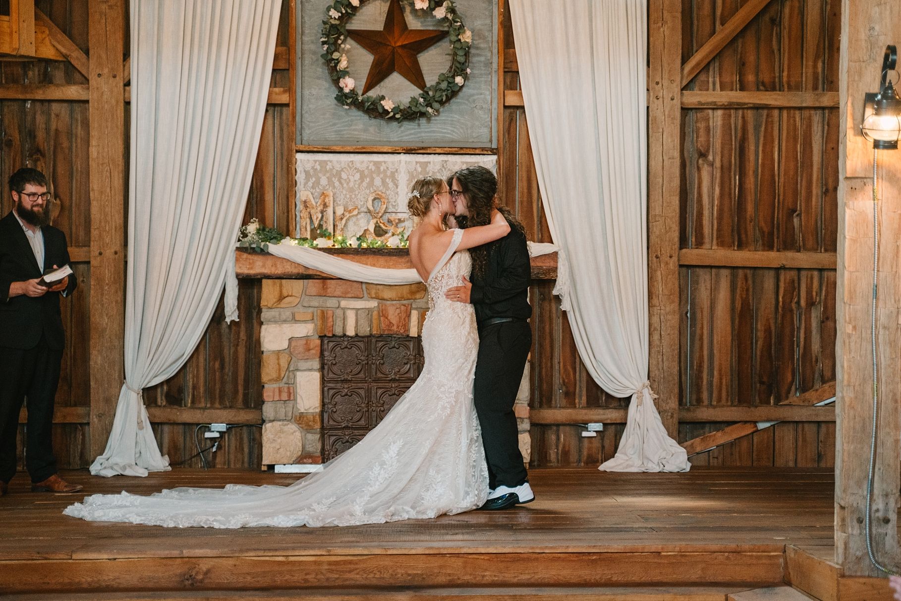 Newlyweds sharing first kiss on wooden stage in decorated rustic barn wedding venue.