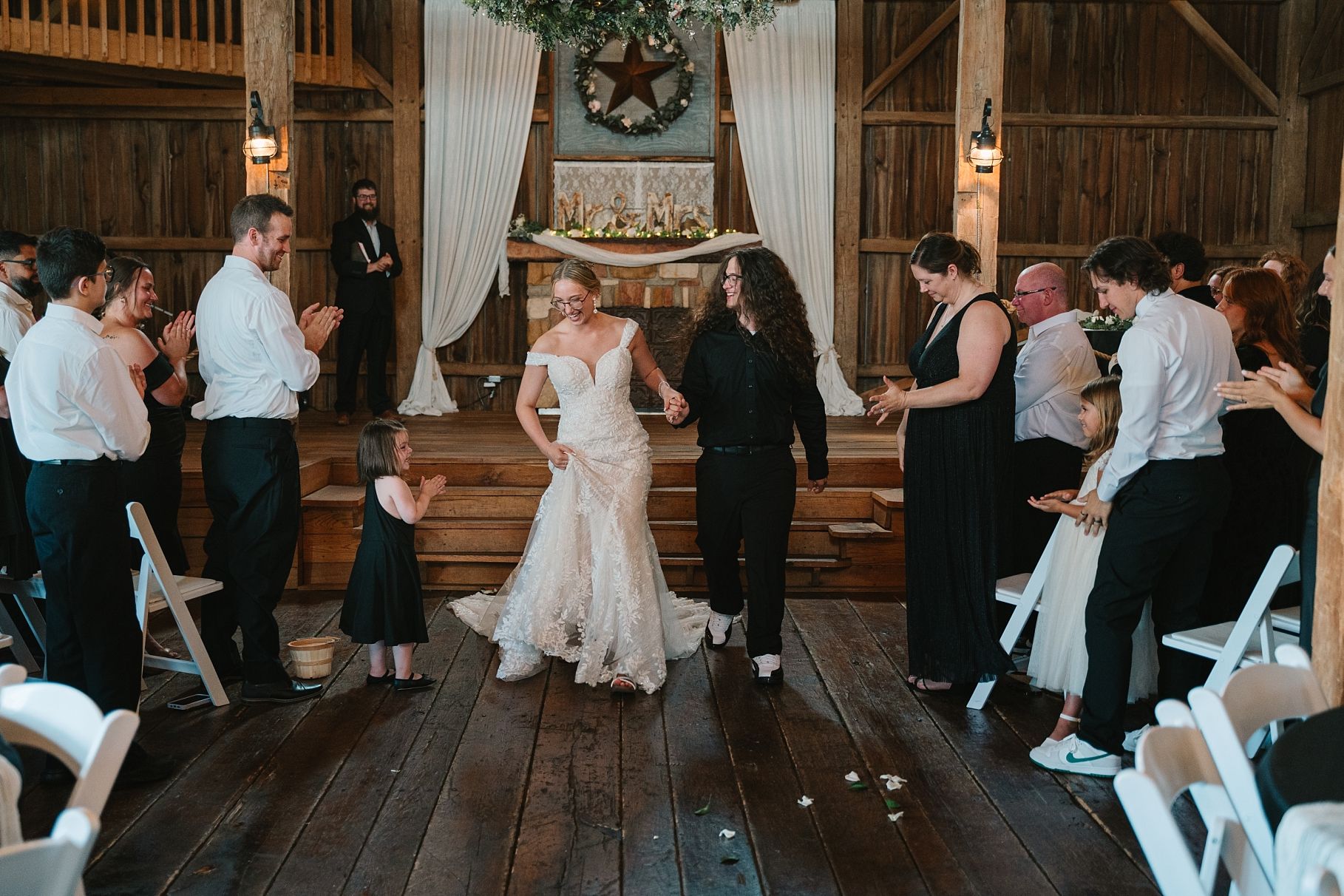 Bride in lace gown walking down aisle with partner through cheering guests in rustic barn venue.