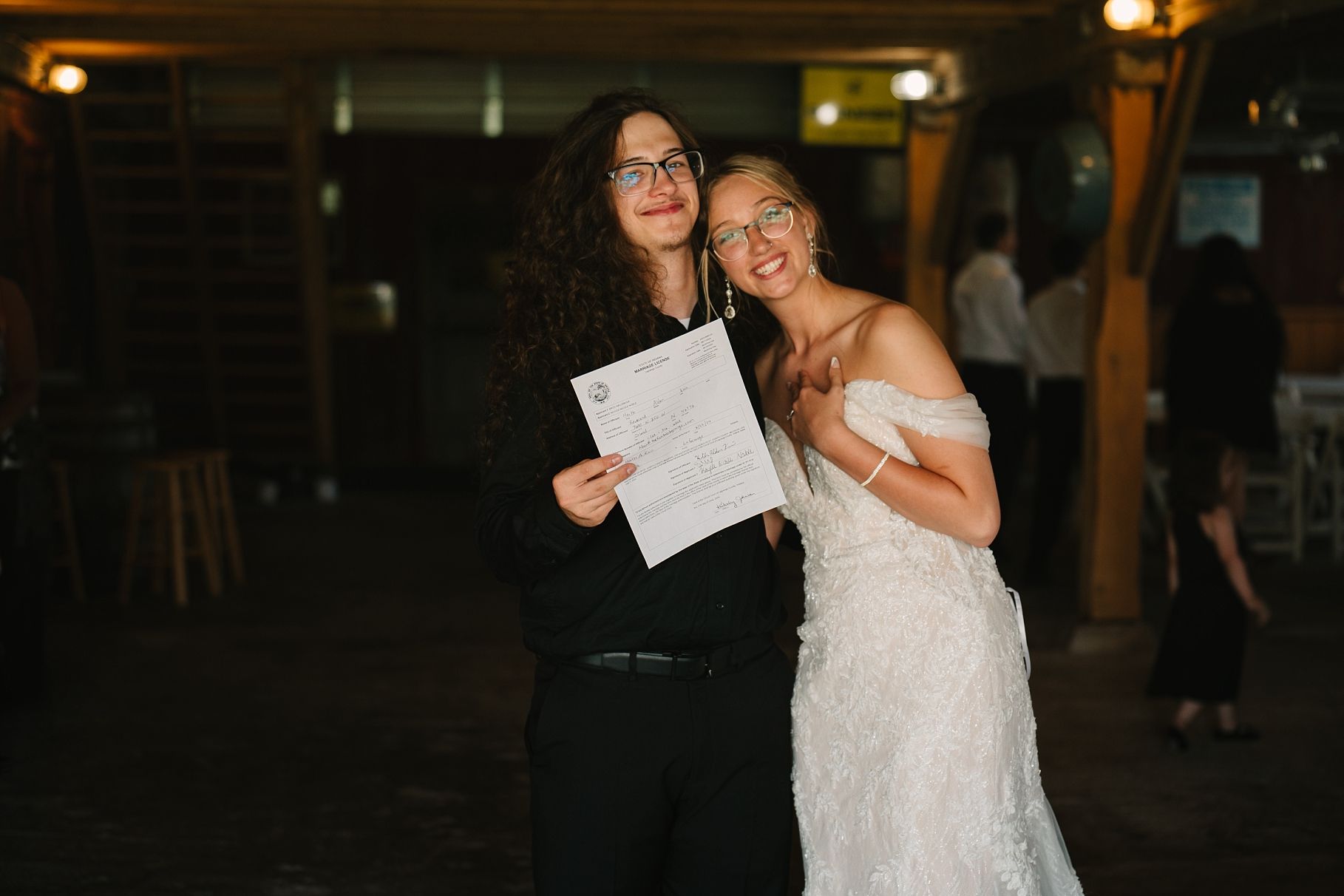 Newlywed couple joyfully posing with marriage certificate after barn wedding ceremony at night.