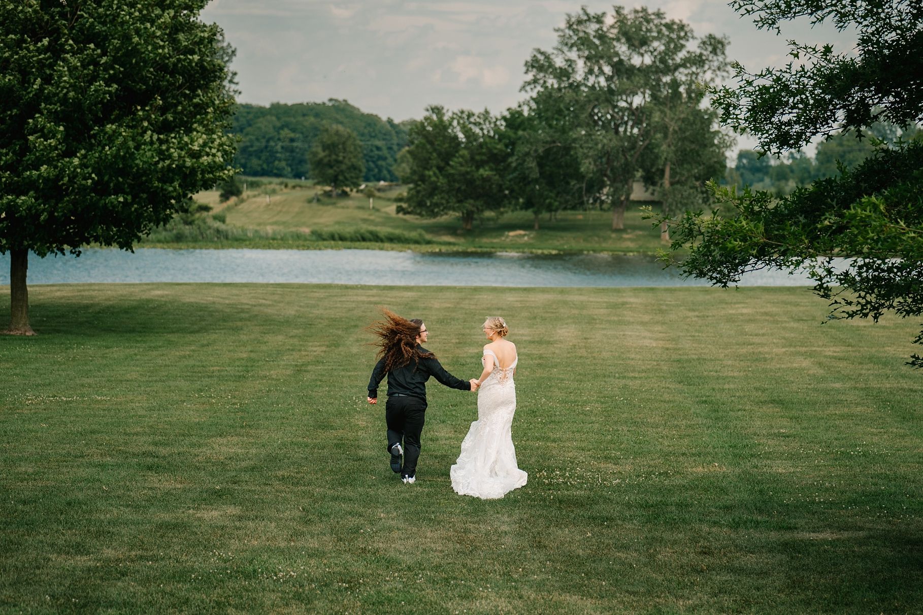 Newlywed couple running hand in hand across green lawn with scenic lake backdrop.