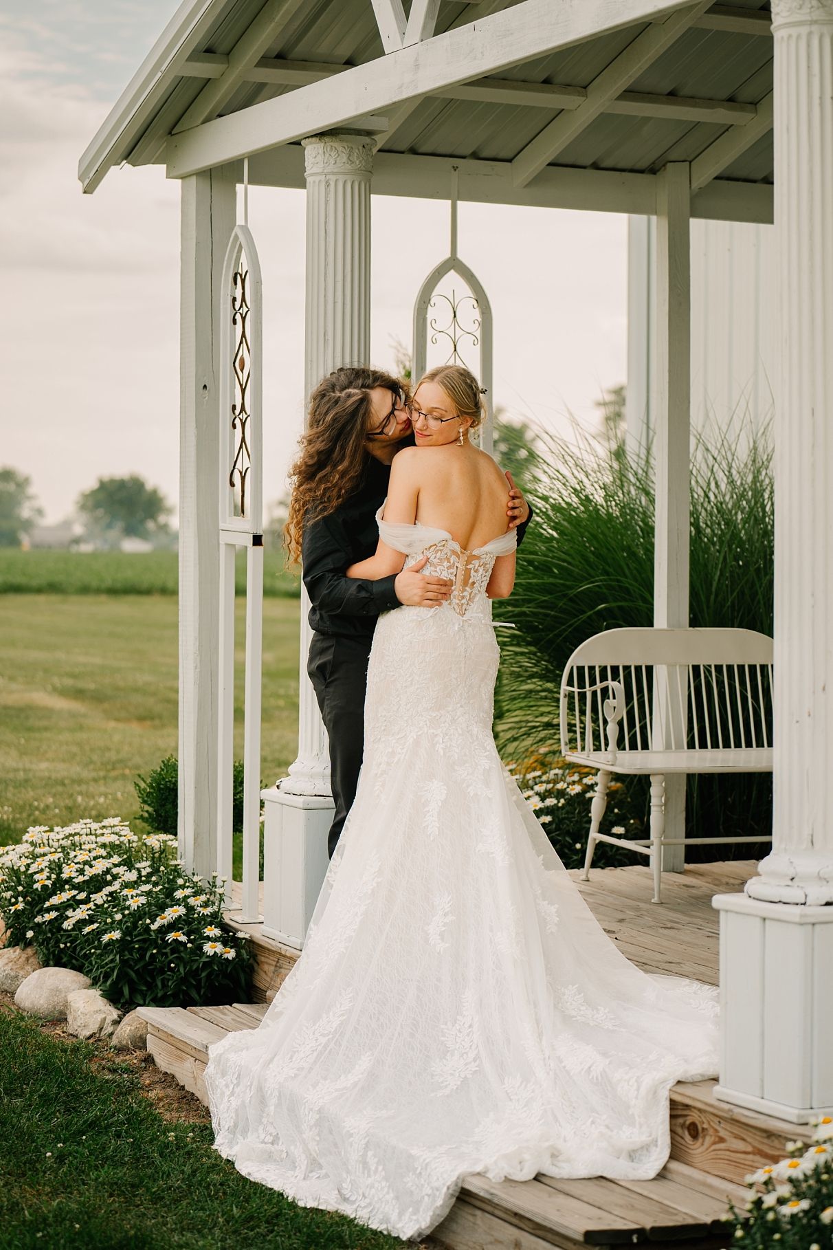 Couple sharing romantic kiss under white gazebo surrounded by wildflowers at golden hour.