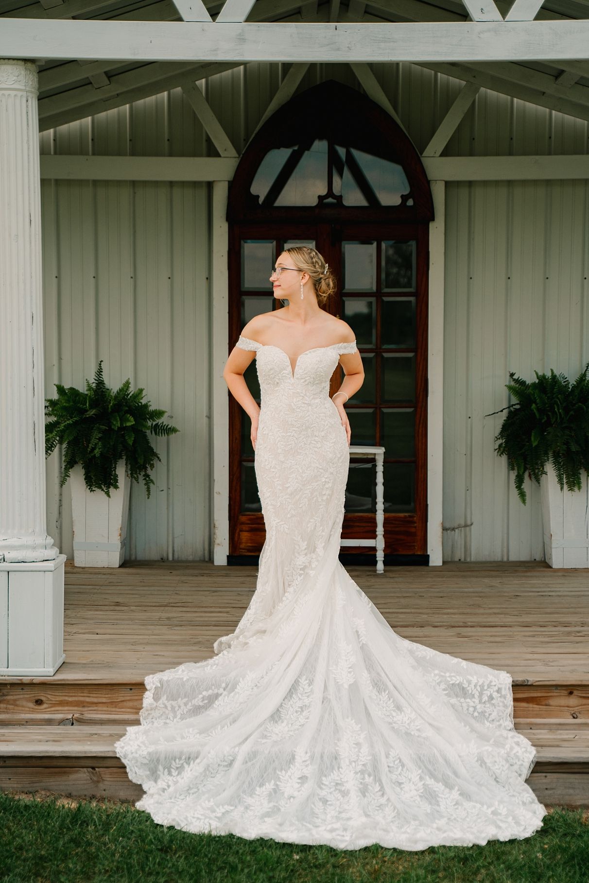 Bride in off-shoulder lace mermaid wedding gown with train poses on white barn venue porch.