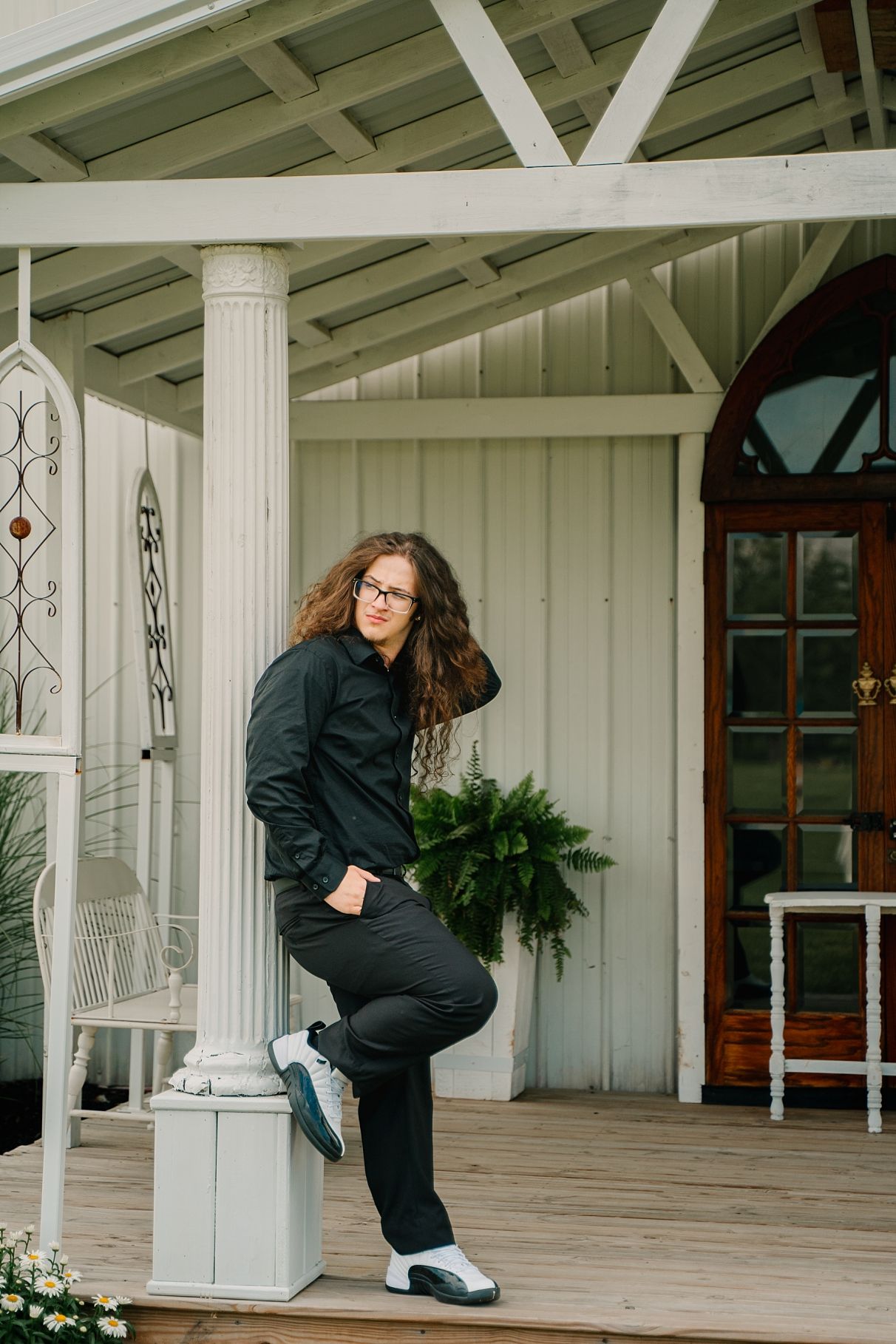 Curly-haired person in all-black outfit leans against white porch column of rustic venue.