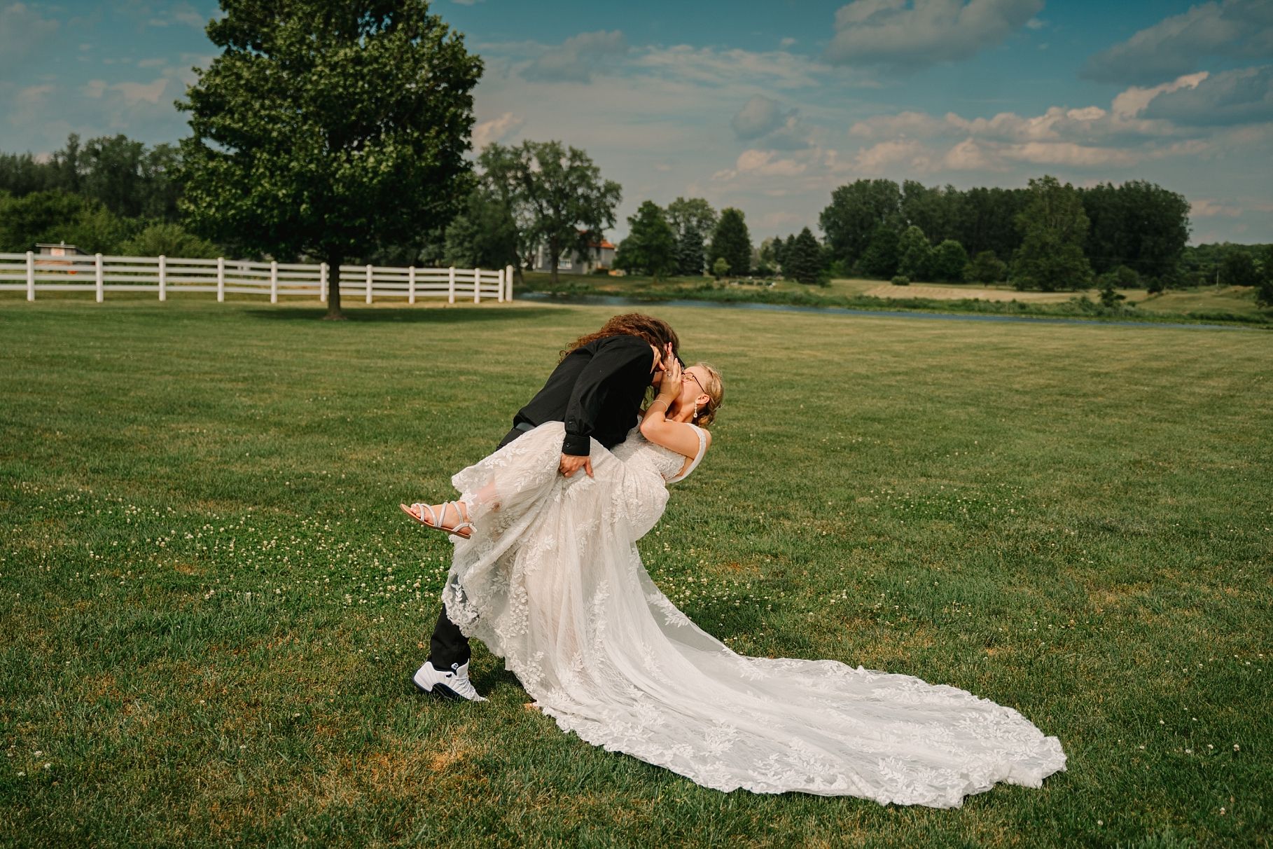 Groom dips bride in white lace wedding dress on lush green lawn at outdoor wedding.