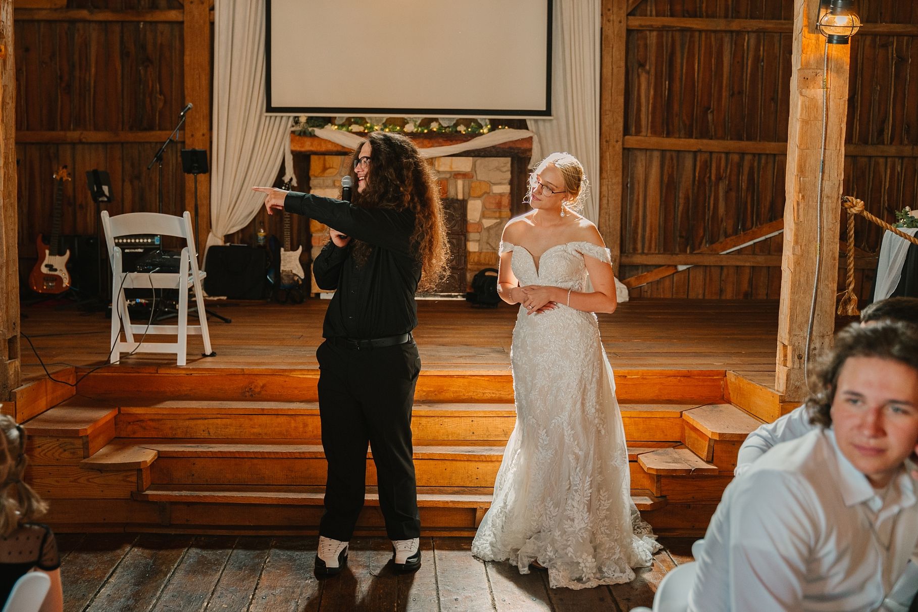 Wedding coordinator adjusts brides off-shoulder lace gown inside rustic barn reception hall.