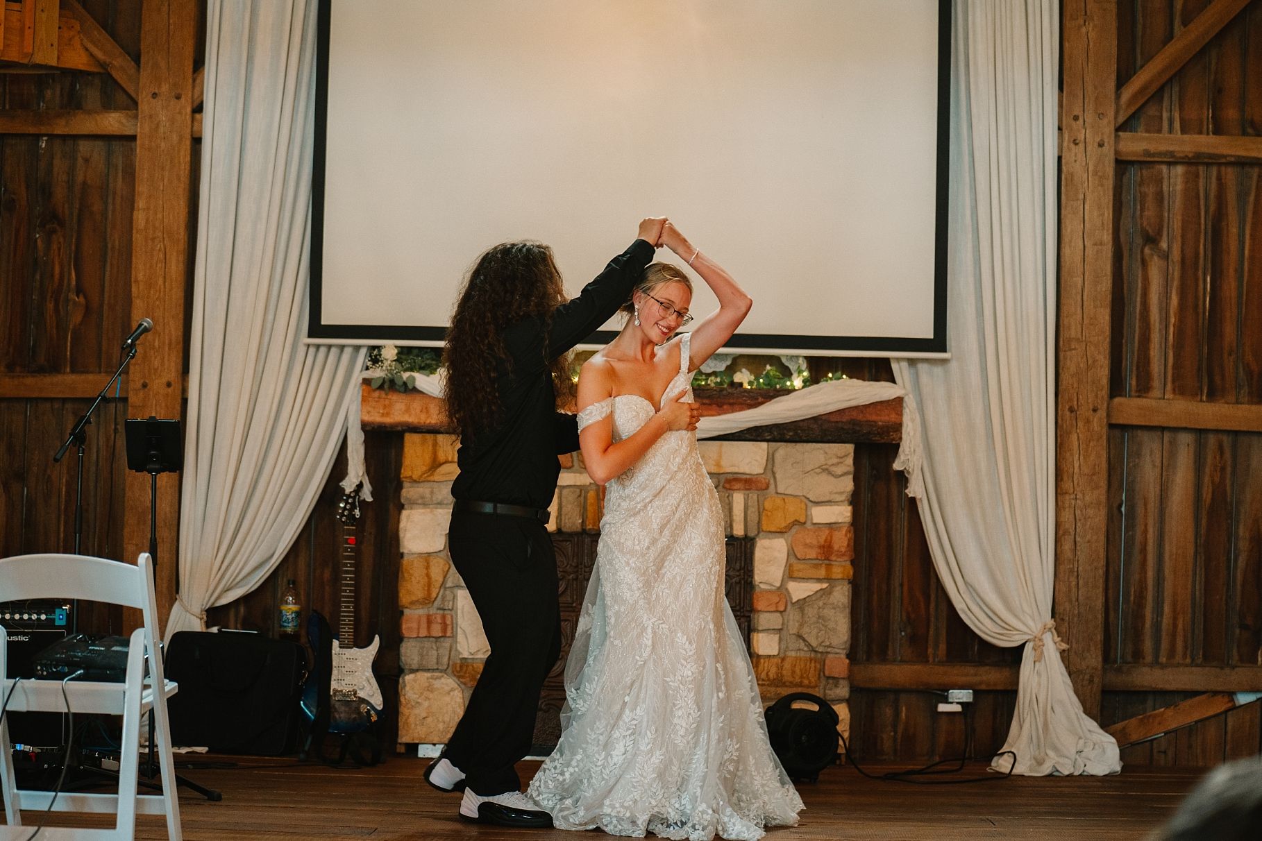 Couple shares first dance spin near stone fireplace in warmly lit rustic barn wedding venue.