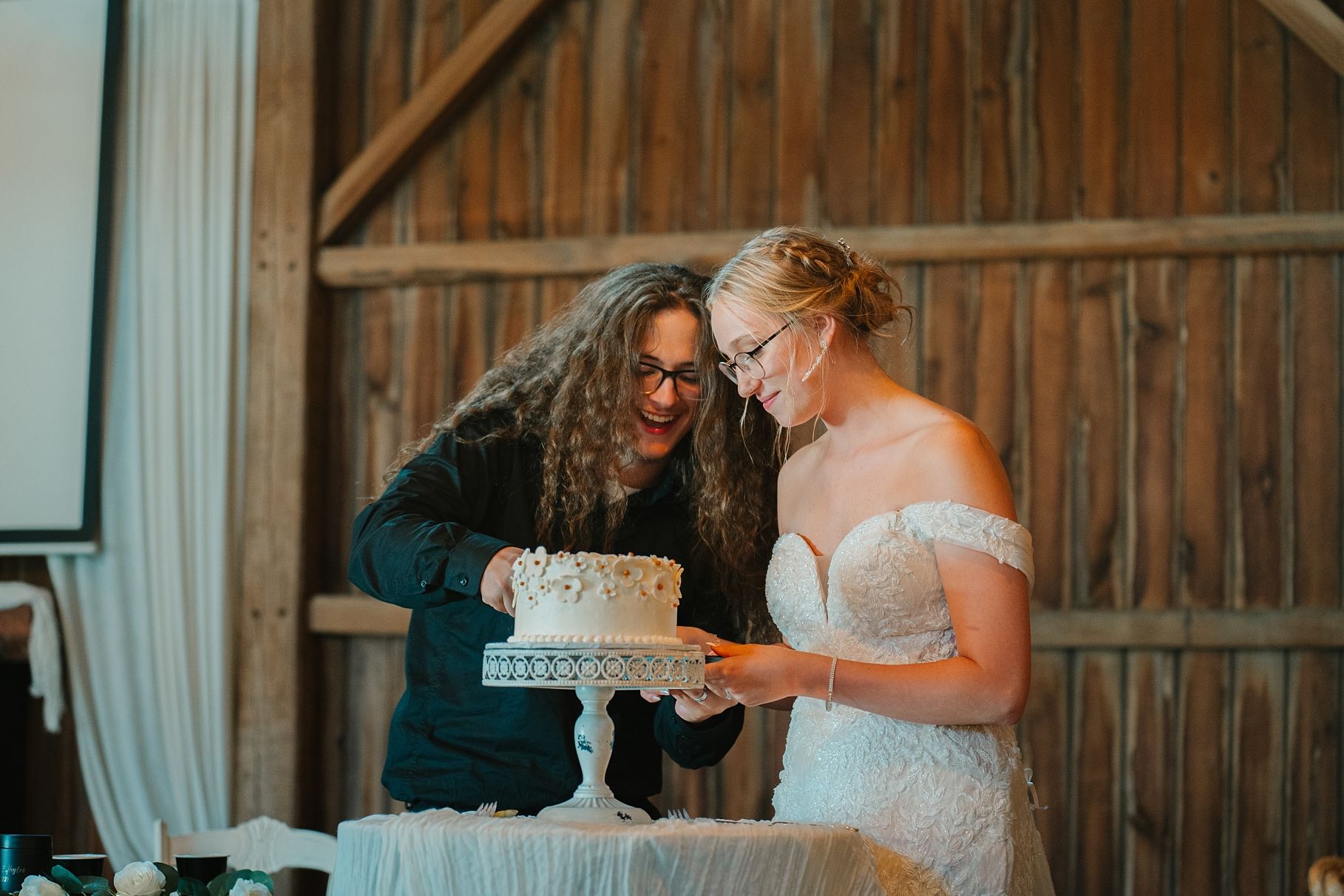 Couple cuts elegant white wedding cake together at rustic barn wedding reception celebration.