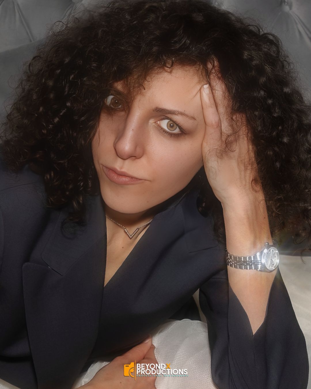 Woman with curly dark hair resting her head on her hand, wearing a navy blazer and silver watch.