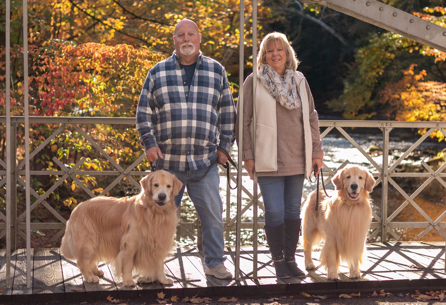 Two golden retrievers sit with their owners on a wooden bridge surrounded by vibrant autumn foliage.