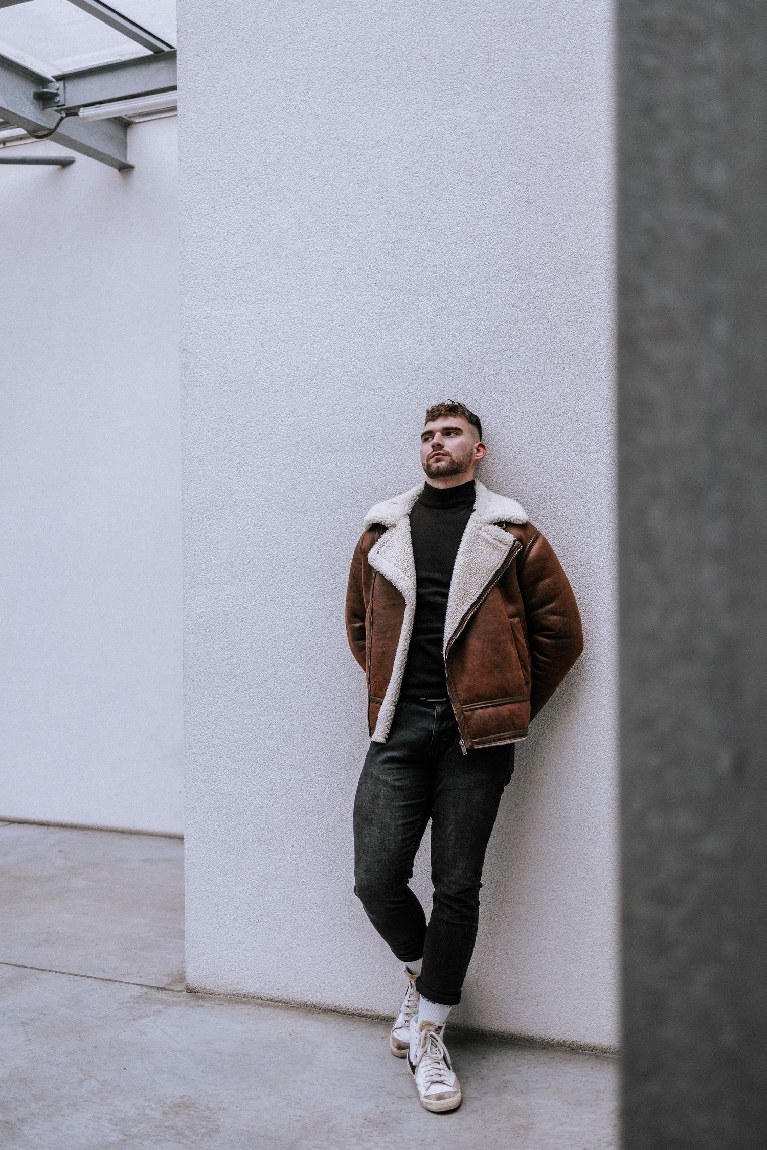 Postava muža v štýlovom zimnom outfite opierajúca sa o modernú budovu. Street style fotografia a mužský portrét, Rajecké Teplice.