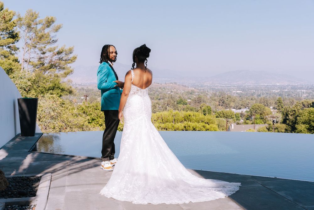 A couple in wedding attire stands on a scenic overlook with mountains and city views in the background.