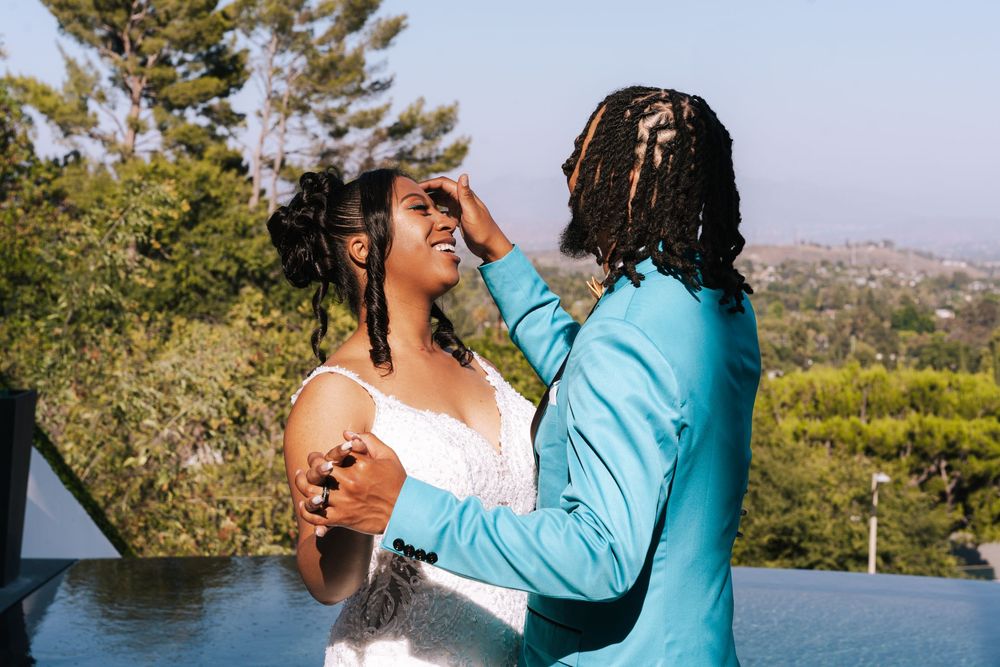 A couple in white and turquoise outfits embrace and laugh together during an outdoor photoshoot in nature.