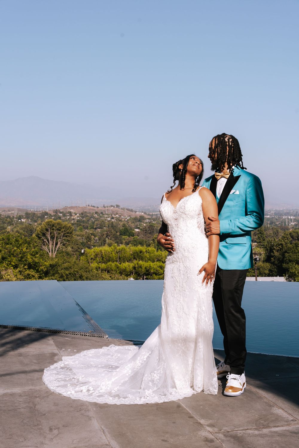 A couple in wedding attire embraces while standing on a scenic overlook with mountains and water in the background.