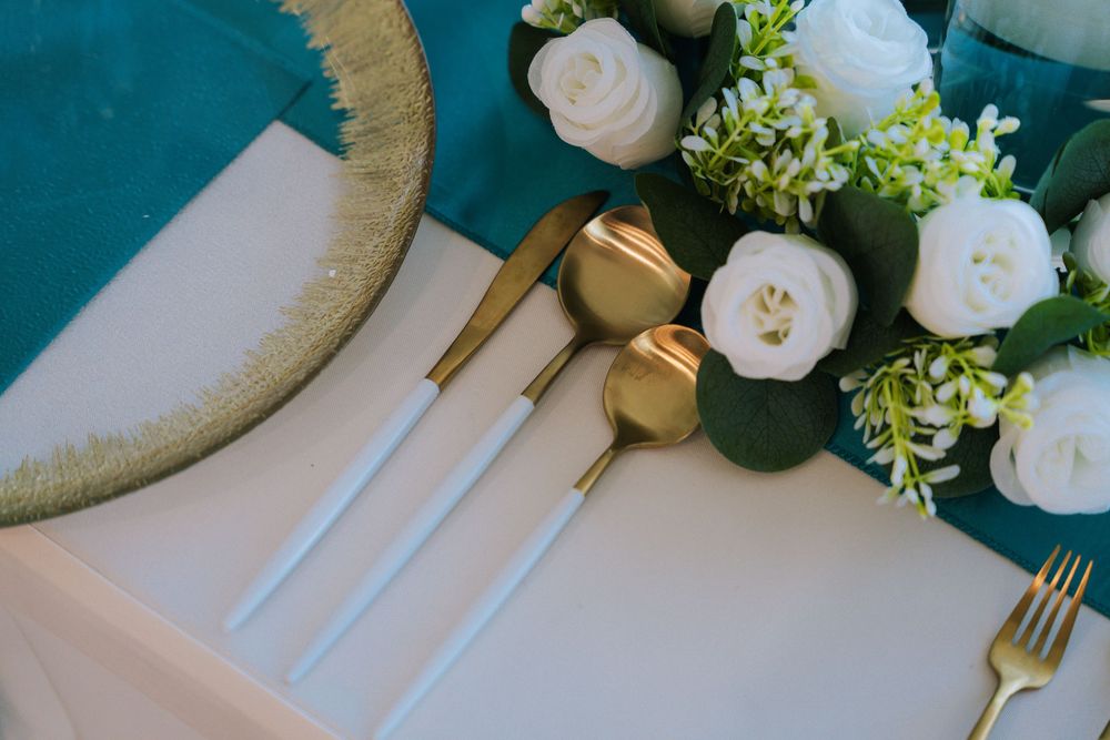 Elegant place setting with white roses, gold cutlery, and eucalyptus on a teal and white background.