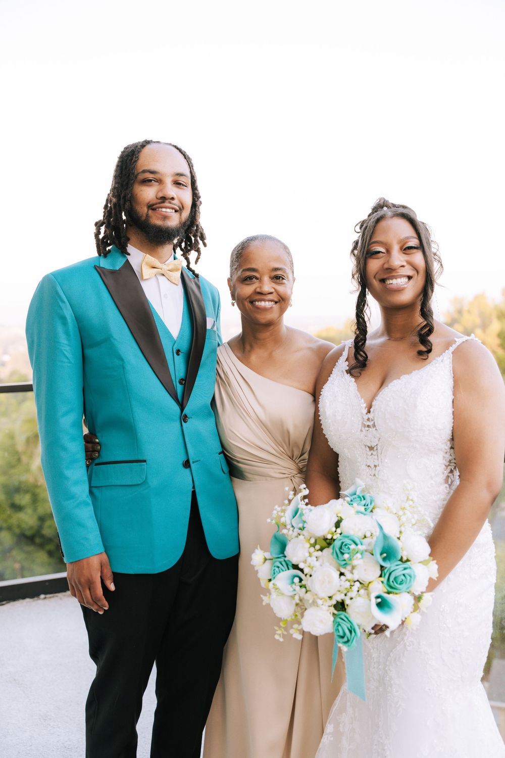 Three people smile together at a wedding celebration, with the groom in a turquoise blazer and bride holding a teal bouquet.