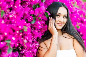 Woman with long dark hair poses against vibrant pink bougainvillea flowers in a garden setting.