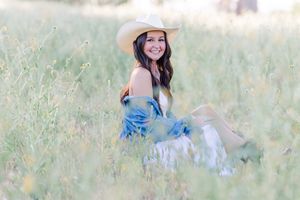 Someone wearing a blue dress and cowboy hat sits in a sunlit meadow of tall grass on a summer day.