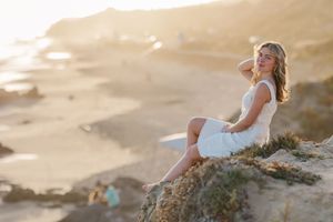 A woman in a white dress walks along a sandy beach at sunset with waves gently rolling in the background.