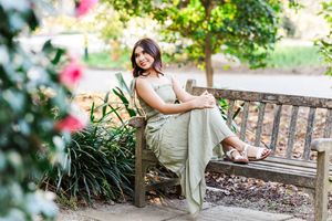 A person in a flowing beige dress sits gracefully on a wooden bench surrounded by lush garden foliage.