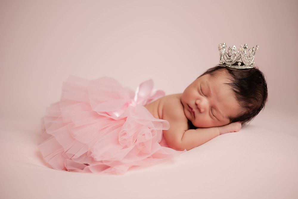 Newborn baby girl sleeping in pink tutu skirt with silver crown on pink background.