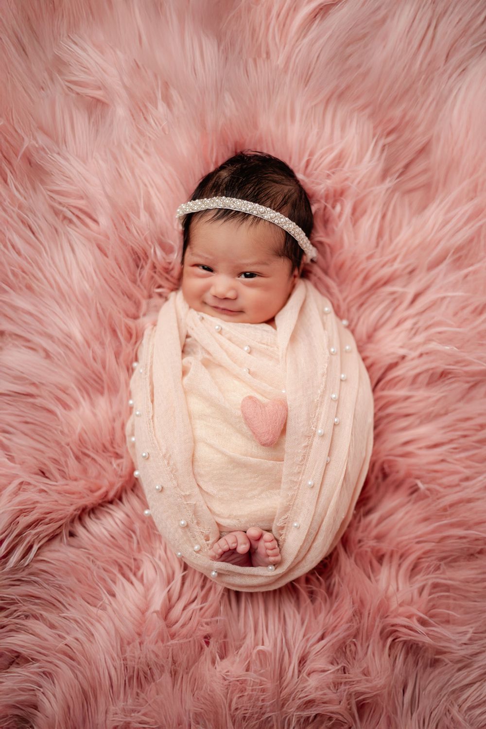 Smiling newborn baby girl swaddled in pink wrap with heart detail, wearing pearl headband on fluffy pink fur.