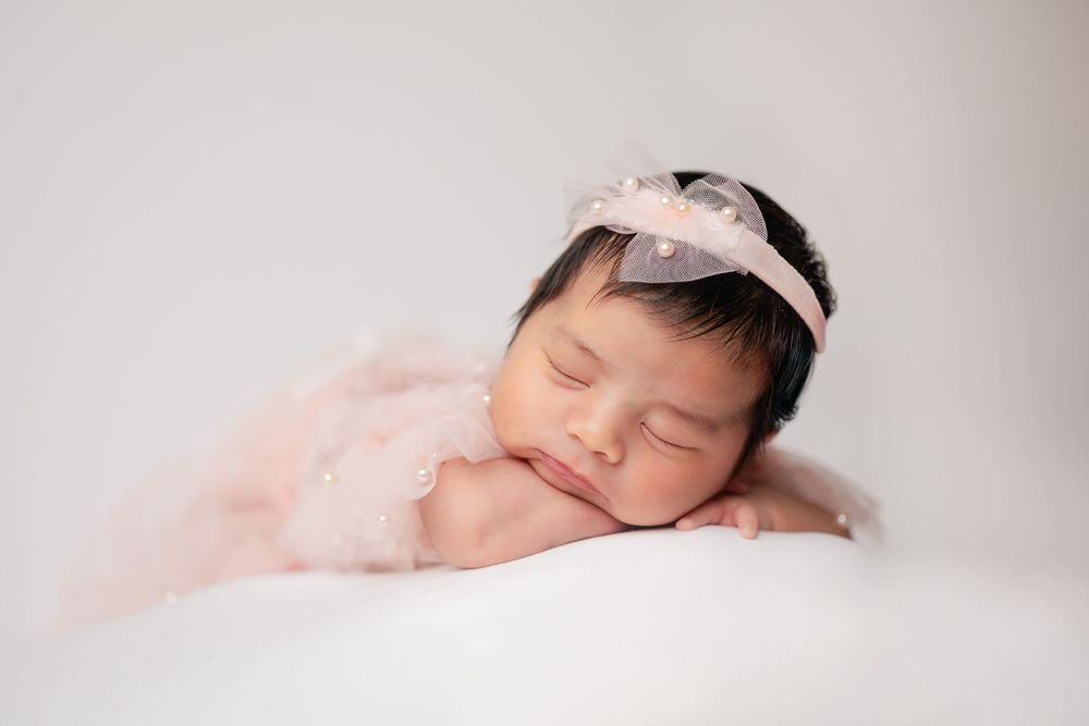 Sleeping newborn baby girl in pink lace outfit with bow headband on white background.