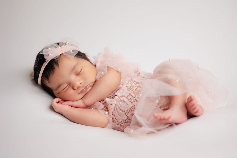 Peaceful newborn baby girl sleeping in pink lace romper with matching headband on white backdrop.