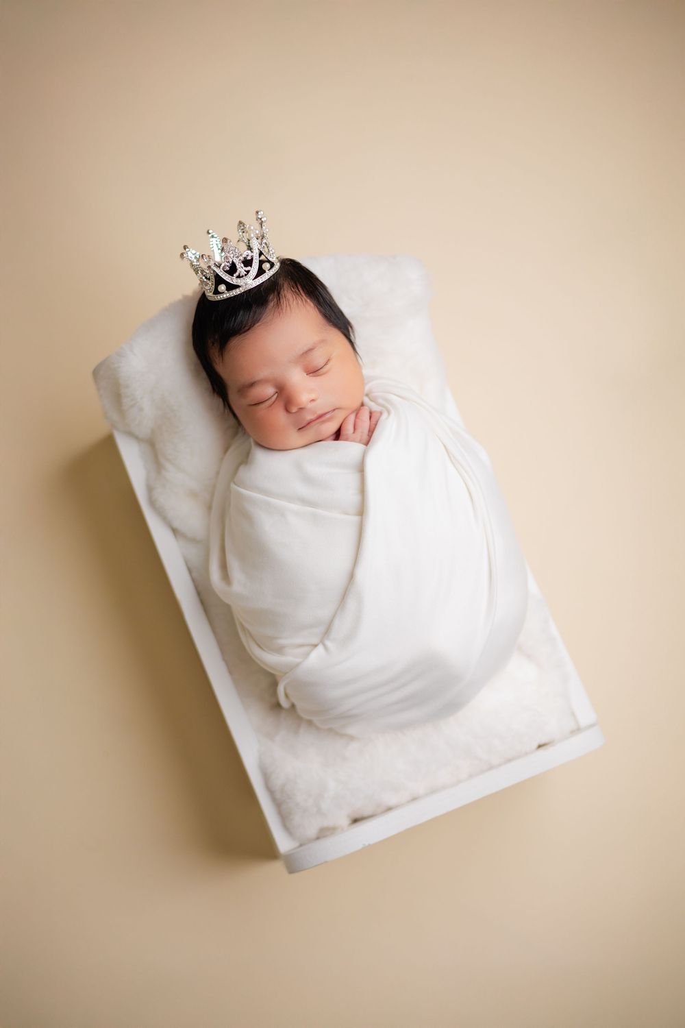 Newborn baby swaddled in white wrap with silver crown in small white bed on beige background.