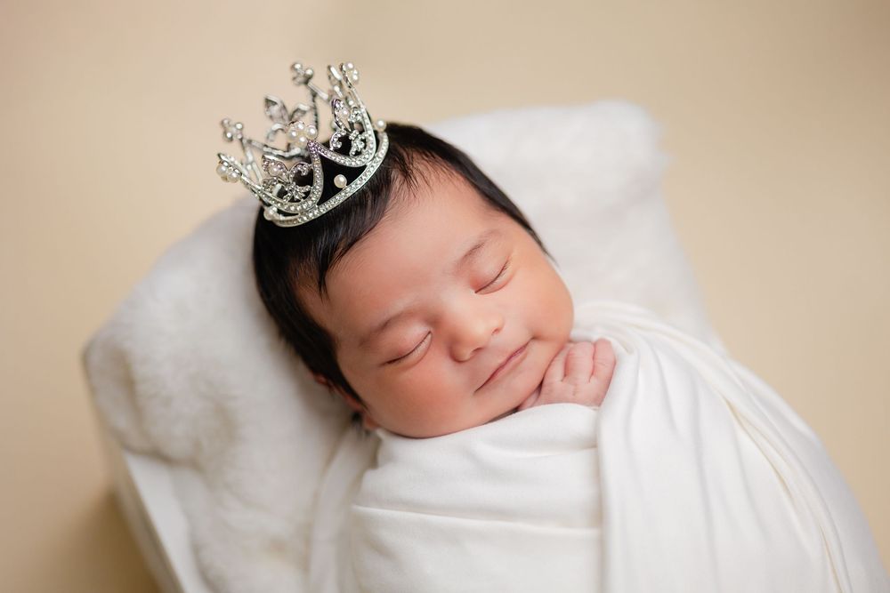 Sleeping newborn baby wearing silver crown swaddled in white wrap on soft white pillow.