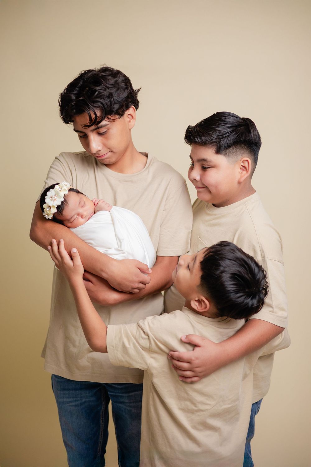 Three brothers in beige shirts and jeans tenderly holding sleeping newborn baby with floral headband.