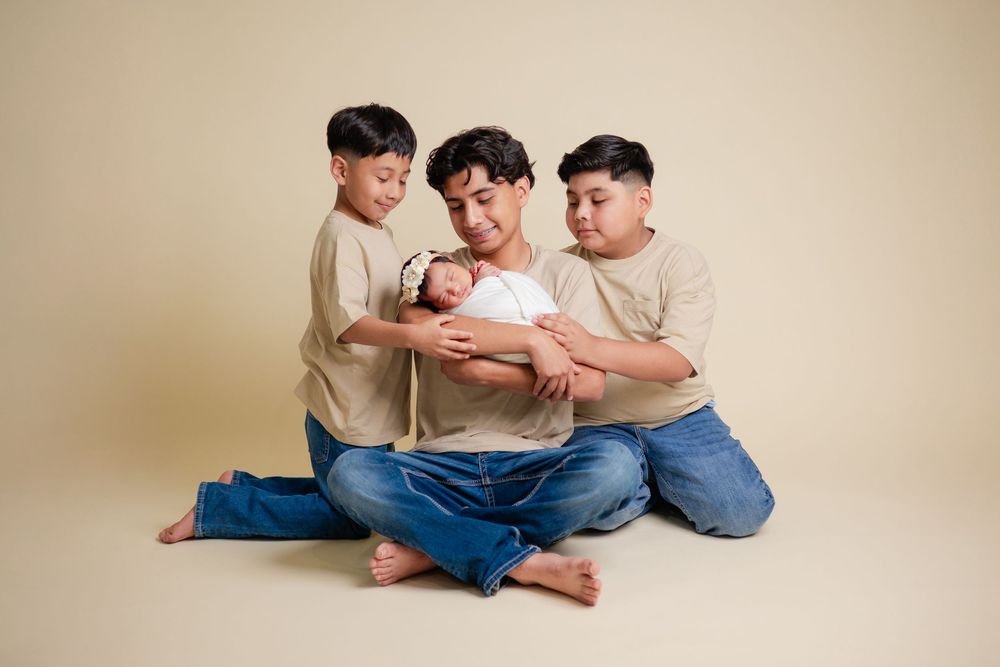 Father sitting on floor holding newborn baby surrounded by two young sons in matching outfits.