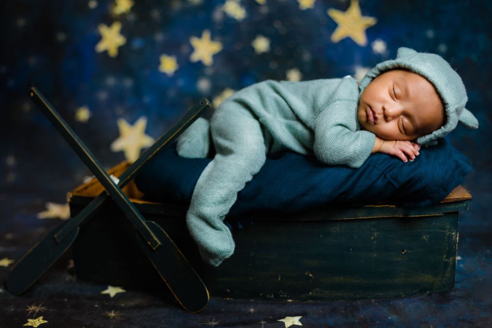 Newborn baby sleeping in vintage suitcase wearing teal knit bear outfit with star backdrop.