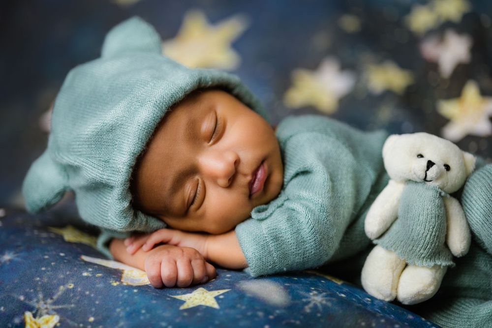Sleeping newborn in teal knit bear outfit beside white teddy bear on starry blue background.