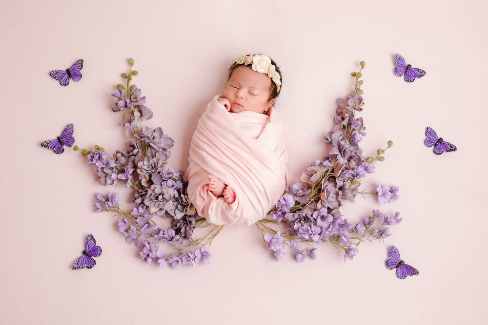 Newborn baby swaddled in pink wrap surrounded by purple lavender flowers and butterfly decorations.